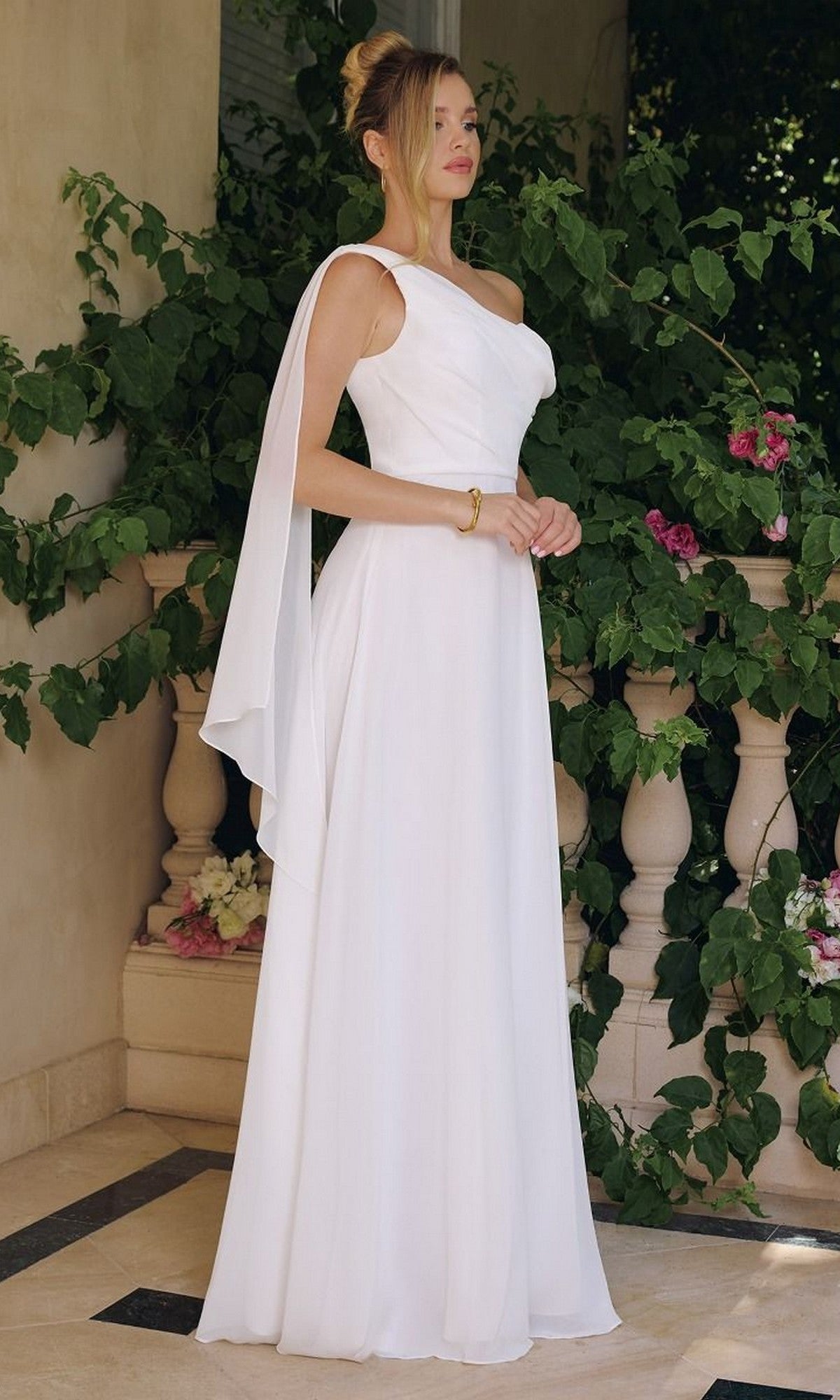 A woman stands outdoors in the Asymmetrical Off-White Bridal Gown: Amoris T716W, featuring a one-shoulder draped sleeve and flowing chiffon skirt, surrounded by greenery and flowers.